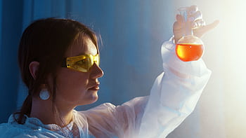 Woman in lab coat examining a vibrant liquid in a flask under lab lights. HD wallpaper