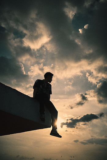 Silhouette of a man sitting on a rooftop edge against a dramatic sunset sky, capturing a moment of solitude and reflection. HD phone wallpaper