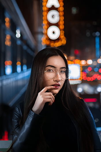 Young woman in glasses posing at night with city lights backdrop and bokeh effect. HD phone wallpaper
