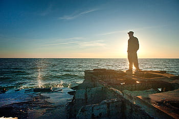 A lone figure stands on a rocky cliff by the ocean during sunset, embracing the serene seascape. HD wallpaper