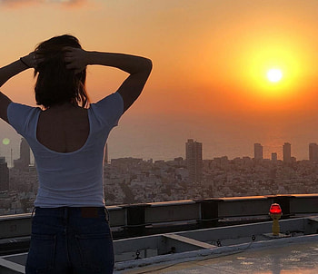 Alona Tal, clasping hands, white t top, brunette, city and ocean below, looking at sunset, jeans HD wallpaper