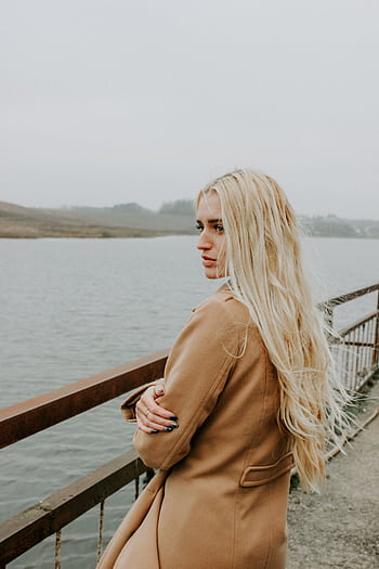 Profile of a woman with long blonde hair in a coat standing on a bridge overlooking a serene lake. HD phone wallpaper