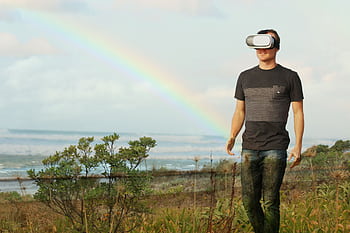 Man using VR headset walks in nature under a rainbow, showcasing modern technology outdoors. HD wallpaper