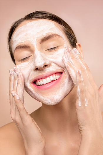 Smiling woman applies facial wash while enjoying her skincare routine, promoting happiness and skin health. HD phone wallpaper