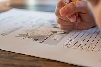 Close-up of a child's hand writing answers on a worksheet in a school setting, emphasizing learning and study. HD wallpaper