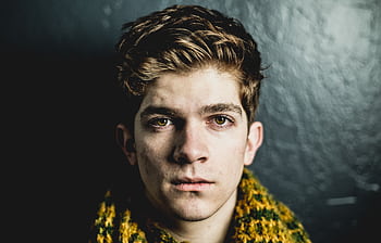 Close-up portrait of a young adult male with a serious expression against a dark textured wall. HD wallpaper