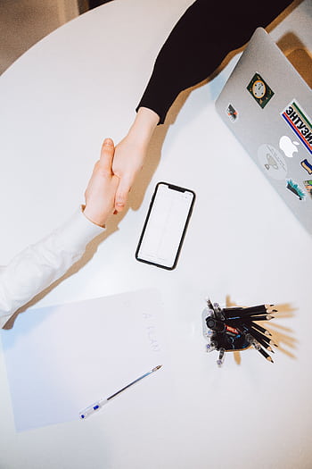 Two individuals shaking hands across a desk with a smart and laptop in a modern office setting. HD phone wallpaper