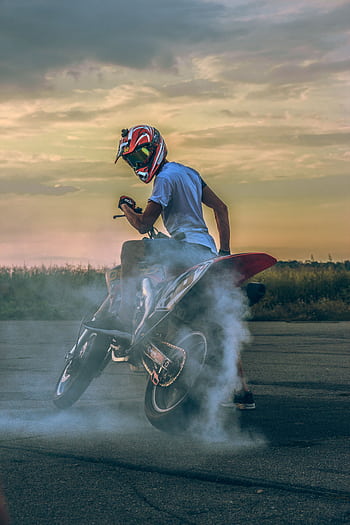 Motorcyclist performing a drift on pavement, creating smoke under a sunset sky. HD phone wallpaper