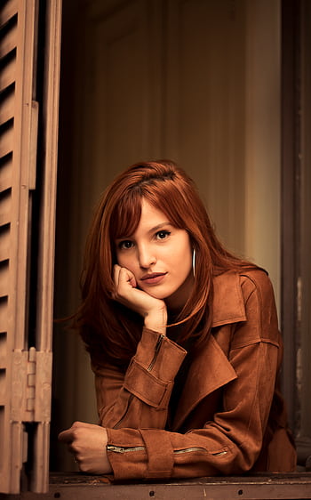 A fashionable redhead woman in a brown jacket poses charmingly by a window indoors. HD phone wallpaper