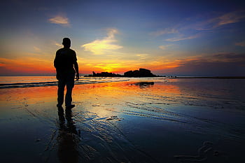 A silhouette of a person stands on the beach reflecting the vibrant sunset sky and tranquil sea. HD wallpaper