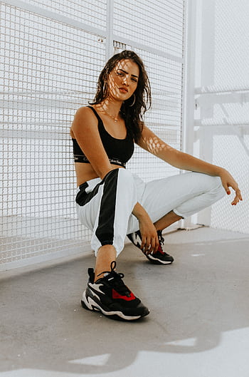Confident woman in urban attire, featuring trendy sneakers, crouching in sunlight. HD phone wallpaper