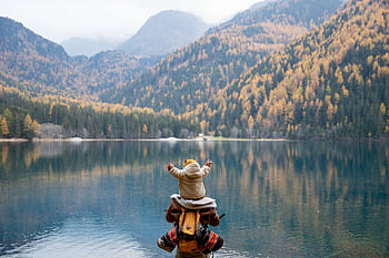 A parent and child seated on a lakeside log, enjoying the serene mountain view in autumn. HD wallpaper