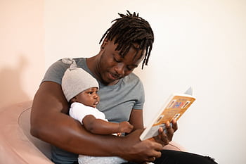 A father with dreadlocks reads a book to his baby, creating a bonding moment. HD wallpaper