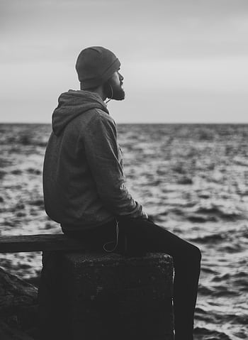 A solitary man sits by the ocean, absorbed in thought, captured in a monochrome scene. HD phone wallpaper