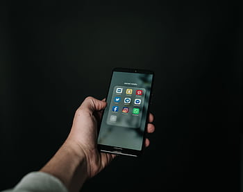 Close-up of a hand holding a smart displaying various social media app icons on a dark background. HD wallpaper