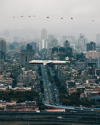 A passenger airplane flying over Taipei's skyline with modern urban buildings and cloudy skies. HD phone wallpaper