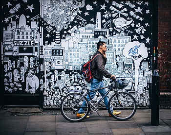 Man walking a bicycle past a detailed urban graffiti mural in a city setting. HD wallpaper
