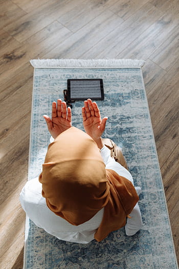 A woman in a hijab prays on a rug with a tablet nearby, in a peaceful indoor setting. HD phone wallpaper