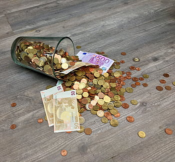 A glass jar with euro coins and banknotes spilled on a wooden surface, representing finance and savings. HD wallpaper