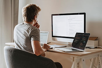 A man deeply engaged in software development with two laptops and a monitor. HD wallpaper