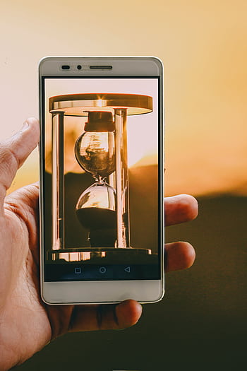 Hand holding smart with hourglass at sunset, symbolizing time and technology. HD phone wallpaper
