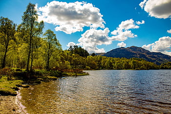 A serene landscape of a lake with surrounding trees and mountains under a clear blue sky. HD wallpaper