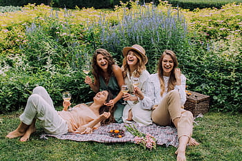 Four women enjoying a picnic, laughter, and wine in a vibrant summer garden. HD wallpaper
