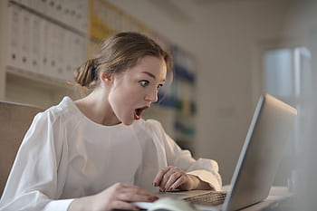 Surprised woman sitting at desk with laptop indoors, expressing amazement. HD wallpaper
