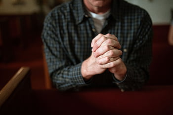 A man in a checked shirt praying in a church, hands clasped together. HD wallpaper
