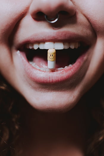 Close-up of a woman with a smiley face pill held between her teeth, evoking themes of medication and wellness. HD phone wallpaper