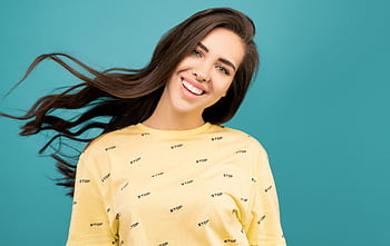 A cheerful woman with flowing hair poses in a yellow shirt against a teal backdrop, exuding happiness and charm. HD wallpaper