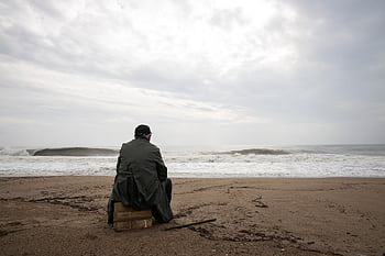 A man sitting alone on an empty beach looking at the stormy sea, evoking thoughts of solitude and reflection. HD wallpaper