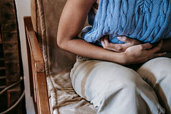 A woman sitting indoors holding a pillow against her stomach, indicating discomfort or pain. HD wallpaper