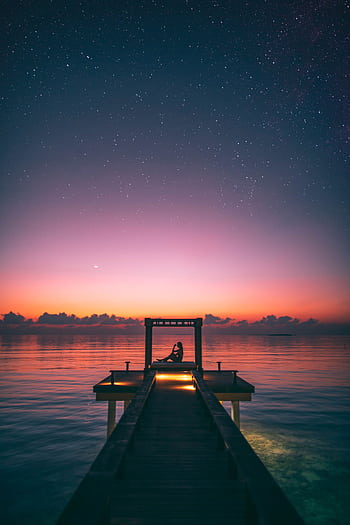Tranquil scene of a person relaxing on a Maldives jetty at twilight under a starry sky. HD phone wallpaper