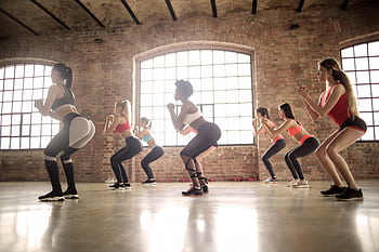 Women participating in a group fitness class performing squats in an industrial-style studio. HD wallpaper