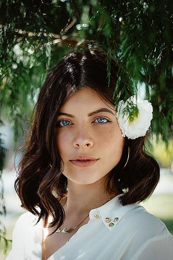 Close-up portrait of a woman with blue eyes and dark hair wearing a white flower outdoors. HD phone wallpaper