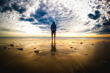 A lone figure stands on a tranquil beach during a vivid sunset, reflecting on the wet sand. HD wallpaper