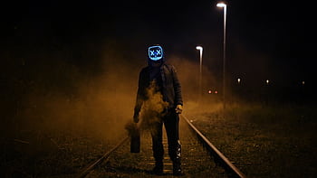 Eerie scene of a person in a neon LED mask standing on railway tracks at night, surrounded by mist. HD wallpaper