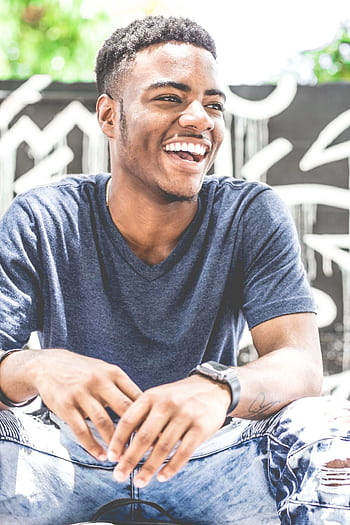Happy young man sitting outdoors with a bright smile, wearing casual clothing. Ideal for lifestyle and fashion themes. HD phone wallpaper