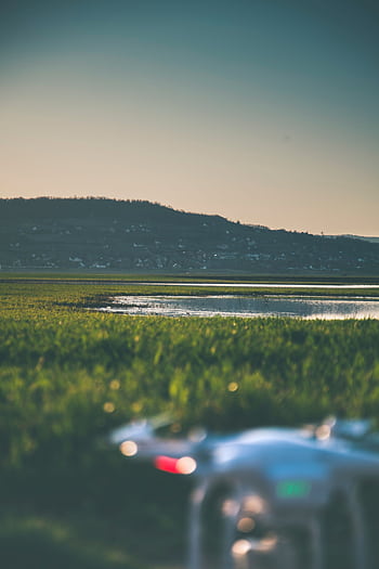 Aerial view of a drone in a green field near a lake with mountains in the background at sunset. HD phone wallpaper