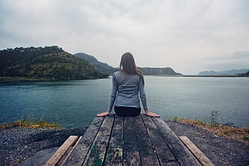 A woman sitting on wooden planks, enjoying a serene mountain lake view. HD wallpaper