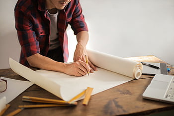 A focused architectural designer drafting blueprints at a desk indoors. HD wallpaper