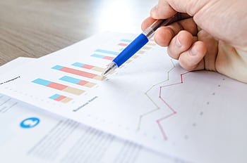 A close-up of a hand with a pen analyzing data on colorful bar and line charts on paper. HD wallpaper