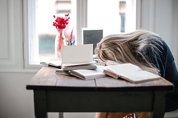 Blonde woman slumped over desk filled with books, showcasing study fatigue indoors. HD wallpaper