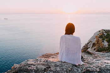 A serene view of a woman sitting on a rocky cliff, gazing at the ocean during sunset. Perfect for relaxation and travel themes. HD wallpaper