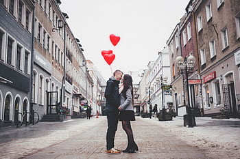 A loving couple shares a kiss with heart-shaped balloons in a charming urban street setting. HD wallpaper