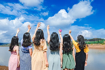 Group of young women with long hair raising hands by a lake under a clear blue sky. HD wallpaper