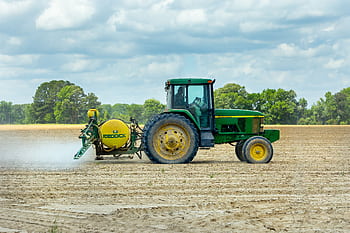 Green tractor with sprayer working in an open farm field under a cloudy sky. HD wallpaper