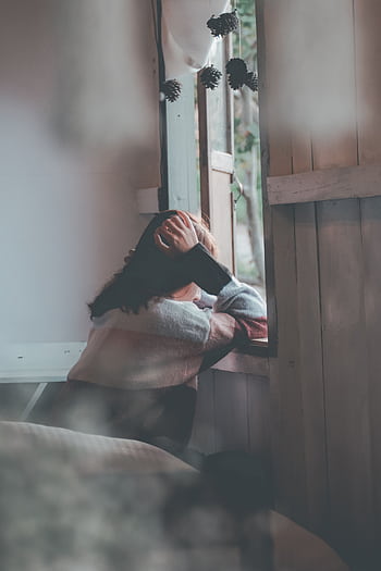 A woman sits indoors gazing thoughtfully out of a window, evoking emotions of loneliness and reflection. HD phone wallpaper