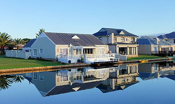 Luxury modern homes reflecting on a peaceful, clear lake during daylight, showcasing architectural beauty. HD wallpaper